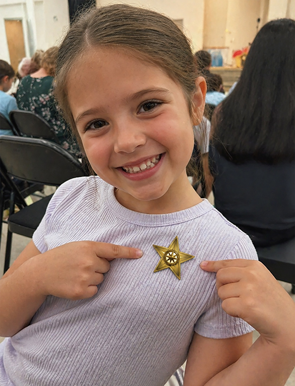 Smiling young girl pointing to her bravery badge from The Garden of Growing Brave, celebrating confidence and courage through the I AM BRAVE Movement.
